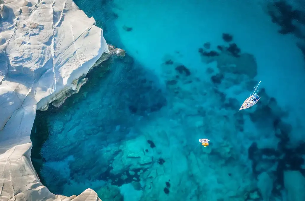 Aerial view of a sailboat and small boat in clear blue water near white rocky coast.